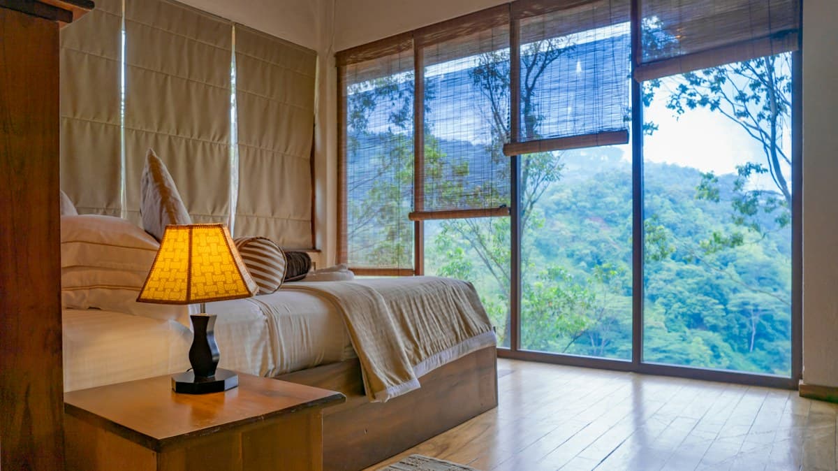 Guest bedroom with white linens, warm wood finishes and large windows facing greenery